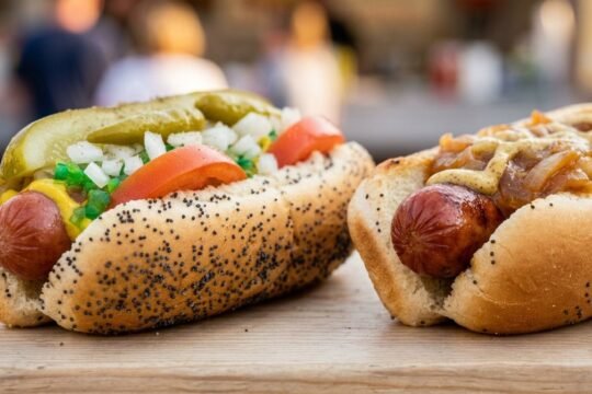 Two hot dogs on poppy seed buns, one Chicago-style and one with caramelized onions