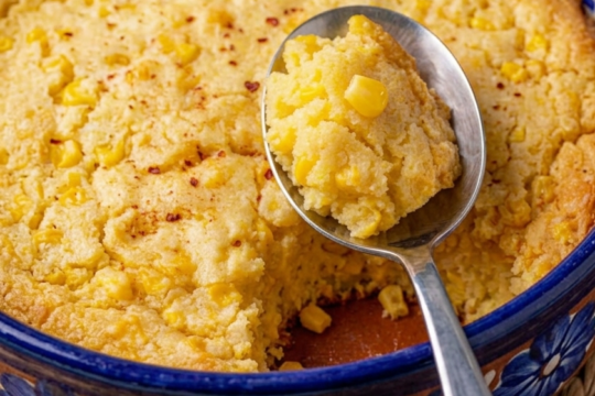 Spoon lifting a golden, corn-studded portion of Jiffy corn casserole from a blue and terracotta ceramic baking dish, dusted lightly with chili powder
