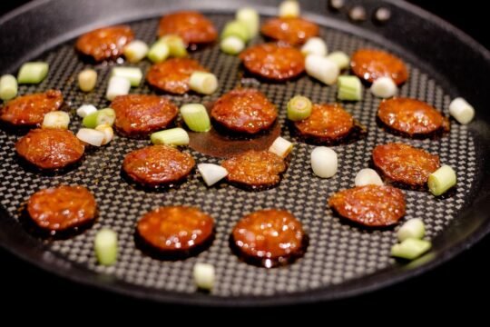 Sliced chorizo fresco pan-frying in a skillet with scallions, showing the deep pimentón-red color