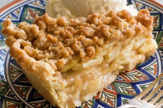 A slice of Dutch apple pie with a golden crumb topping and glossy cinnamon-apple filling, served with a scoop of vanilla ice cream on a decorative plate