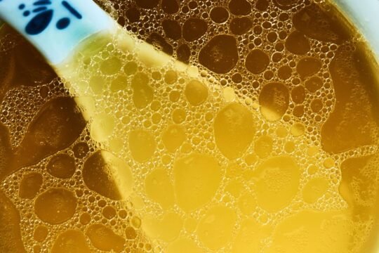 Overhead close-up of clear, golden homemade chicken stock in a white bowl, small beads of chicken fat floating on the surface and the handle of a blue-and-white ceramic spoon resting in the broth
