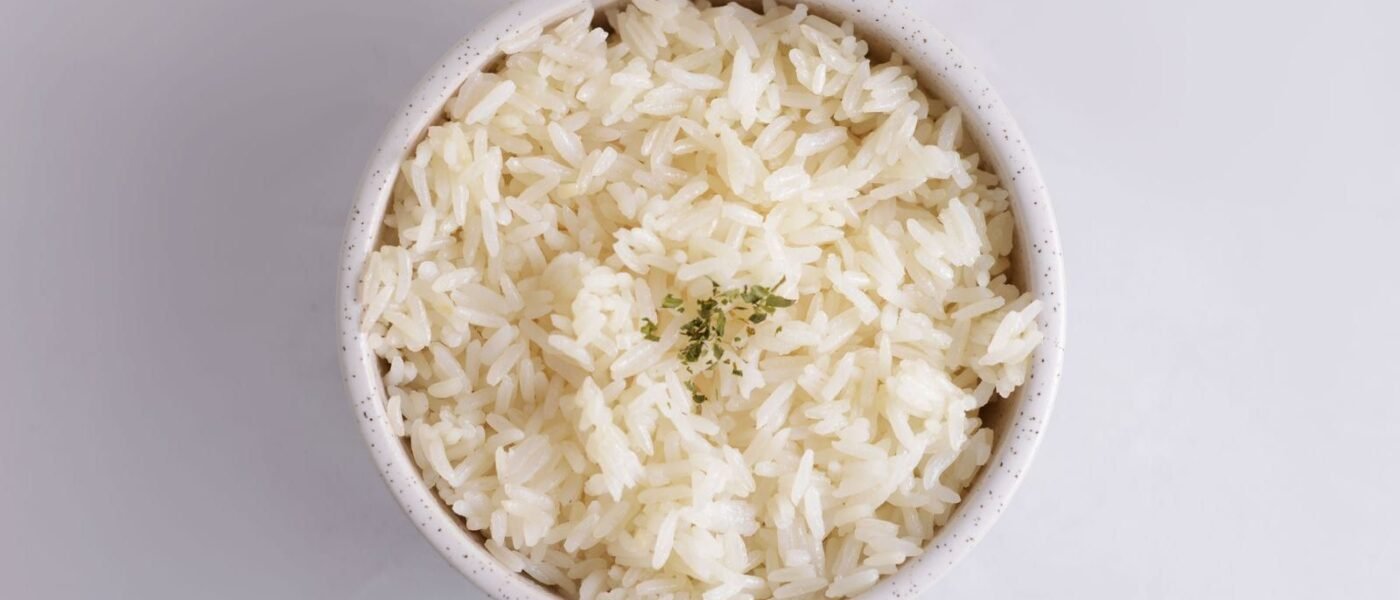 Overhead view of fluffy, separate long-grain white rice in a speckled white bowl, topped with a small pinch of dried herbs, on a pale gray background