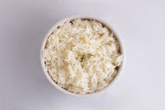 Overhead view of fluffy, separate long-grain white rice in a speckled white bowl, topped with a small pinch of dried herbs, on a pale gray background