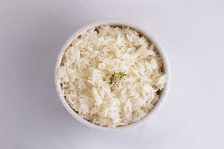 Overhead view of fluffy, separate long-grain white rice in a speckled white bowl, topped with a small pinch of dried herbs, on a pale gray background