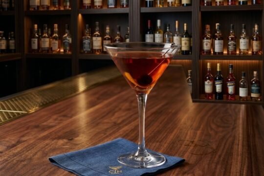 A deep amber Manhattan cocktail in a martini glass, garnished with a brandied cherry and orange twist on a dark wood bar