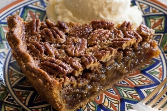 A slice of pecan pie with a glossy caramel filling and whole toasted pecan halves on top, served with a scoop of vanilla ice cream on a decorative plate