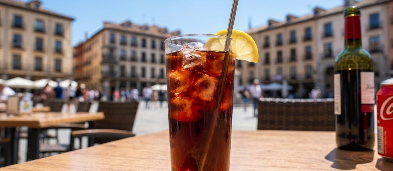 A tall glass of kalimotxo, red wine and cola over ice with a lemon wedge and a metal straw, on a wooden café table in a sunny Spanish plaza, with a bottle of red wine and a can of cola beside it