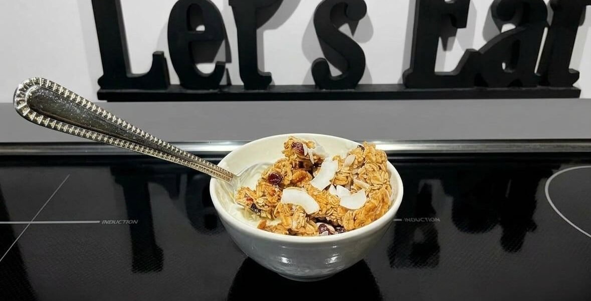 A small white bowl of yogurt topped with golden oat granola clusters, toasted coconut flakes, chia seeds, and dried cranberries, with an ornate silver spoon, sitting on a black induction cooktop in front of a black wooden Let’s Eat sign