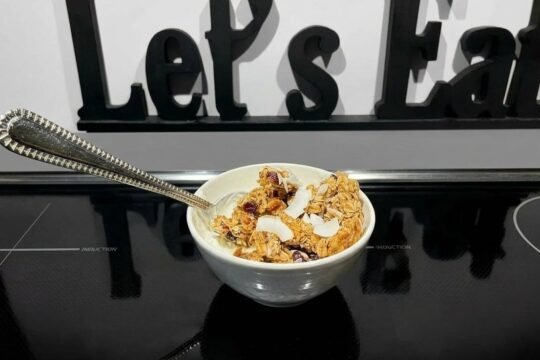 A small white bowl of yogurt topped with golden oat granola clusters, toasted coconut flakes, chia seeds, and dried cranberries, with an ornate silver spoon, sitting on a black induction cooktop in front of a black wooden Let’s Eat sign