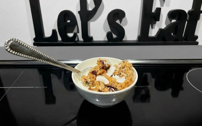 A small white bowl of yogurt topped with golden oat granola clusters, toasted coconut flakes, chia seeds, and dried cranberries, with an ornate silver spoon, sitting on a black induction cooktop in front of a black wooden Let’s Eat sign